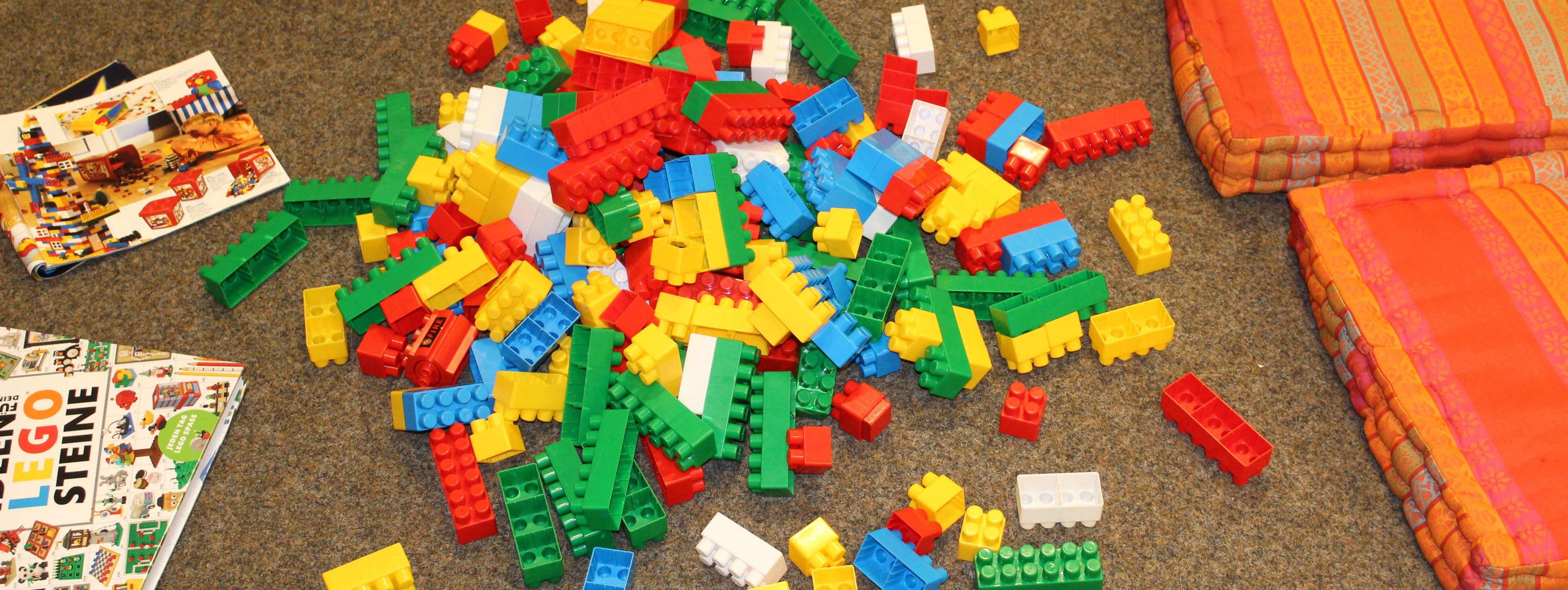 Auf dem braunen Teppich liegen die bunten Lego-Duplo-Steine, drumherum liegen Sitzkissen, auf denen die großen und kleinen Baumeister platznehmen können.