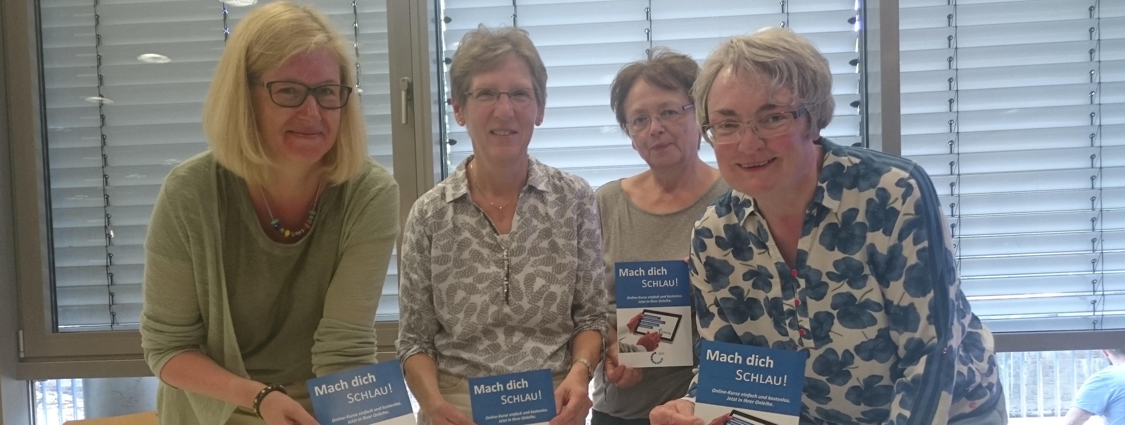 Die Mitarbeiterinnen der Bücherei stehen hinter einem aufgeklappten Laptop und halten lächelnd Broschüren zum E-Learning in Händen