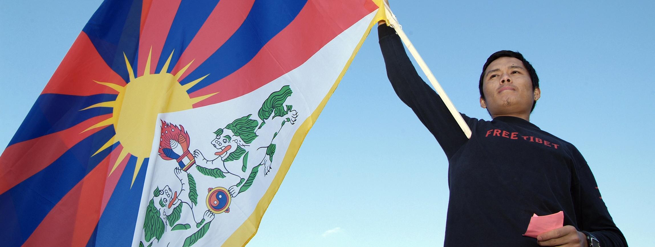 Ein schwarz gekleideter Mann schwenkt die Flagge Tibets. Auf der Mitte der Falgge ist eine Sonne; von ihr gehen sechs rote und sechs blaue Strahlen ab; vor der Sonne ist ein schneebedeckter Berg symbolisch dargestellt; davor sind zwei Seelöwen und ein symbolisches Juwel dargestellt. Die gesamte Flagge ist goldgelb umrandet.