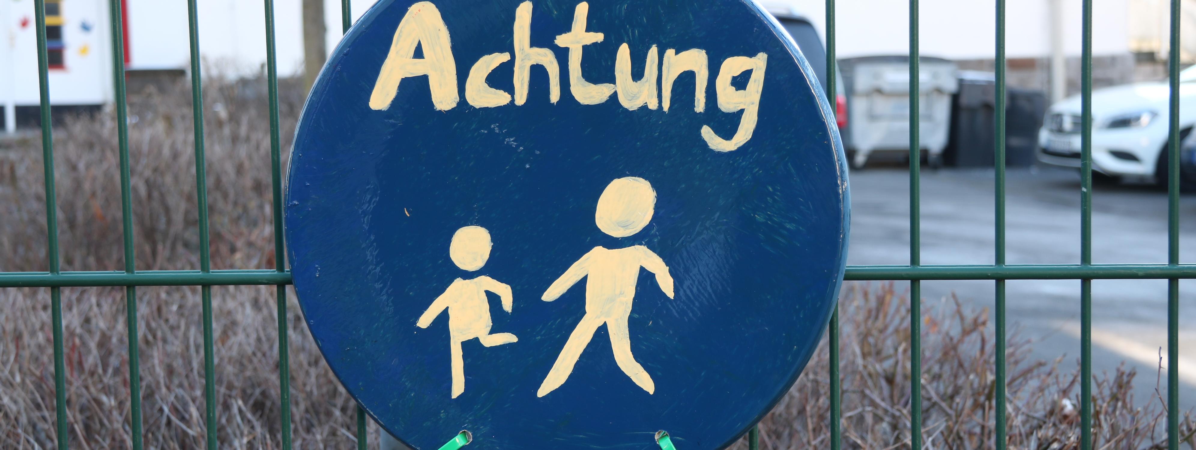 An einem grünen Metallzaun vor der Josefschule hängt ein selbstgemaltes Schild. Es ist rund, darauf sind zwei laufende Strichmännchen in weißer Farbe, darüber steht das Wort "Achtung".