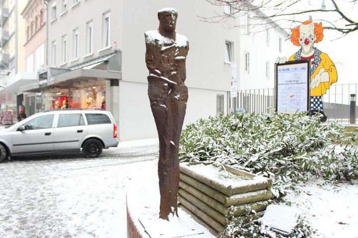 Eine rötliche. aufrecht stehende Skulptur im Winter fotografiert. Auf Kopf und Schultern der Skulptur liegt Schnee. 