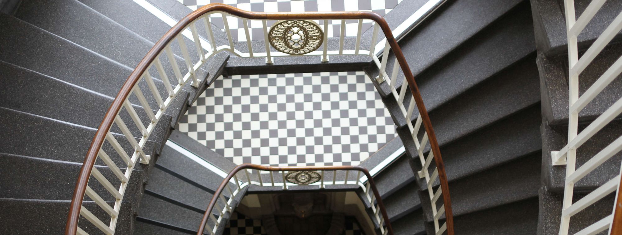 Historisches, oval gehaltenes Treppenhaus, Blick von oben auf weißes Geländer und schwarz weiße Fliesen