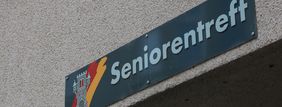 An der Hauswand des Treffs ist ein grünes Schild angebracht, auf dem in weißer Schrift "Seniorentreff" zu lesen ist. Auf dem Schild ist außerdem das Wappen der Stadt Menden zu sehen.