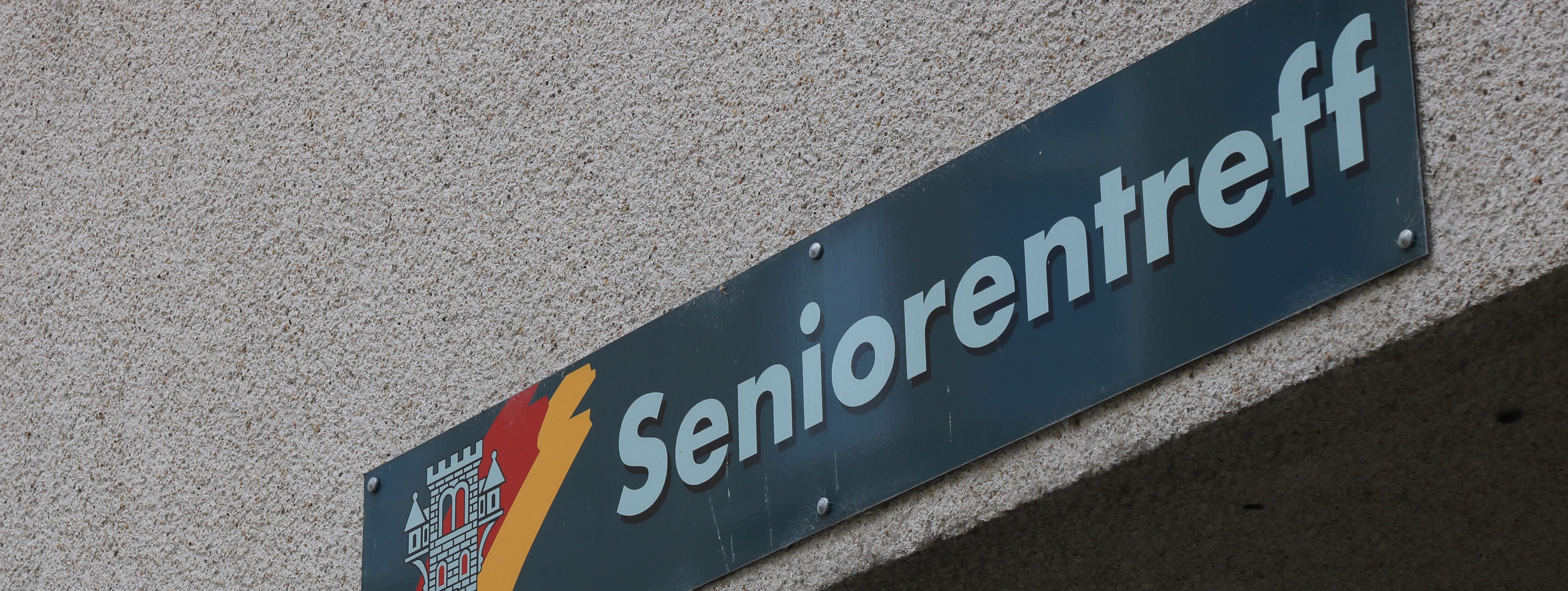 An der Hauswand des Treffs ist ein grünes Schild angebracht, auf dem in weißer Schrift "Seniorentreff" zu lesen ist. Auf dem Schild ist außerdem das Wappen der Stadt Menden zu sehen.