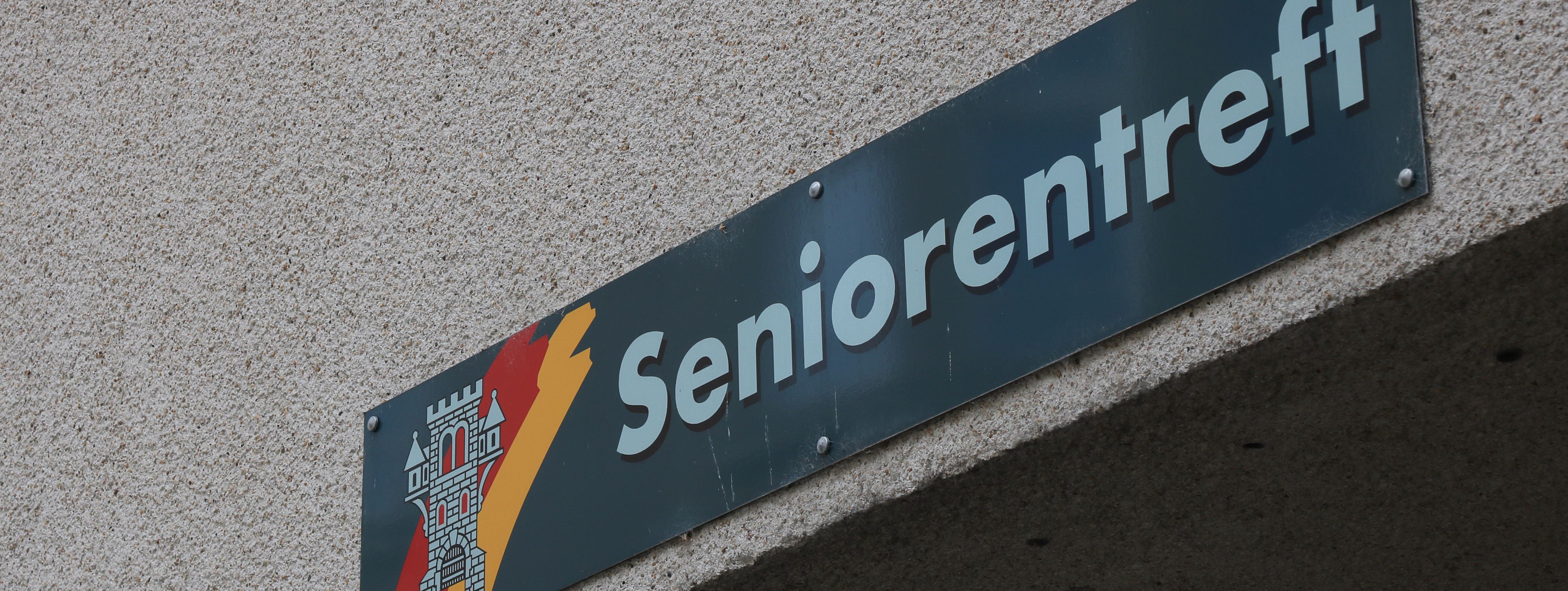 An der Hauswand des Treffs ist ein grünes Schild angebracht, auf dem in weißer Schrift "Seniorentreff" zu lesen ist. Auf dem Schild ist außerdem das Wappen der Stadt Menden zu sehen.