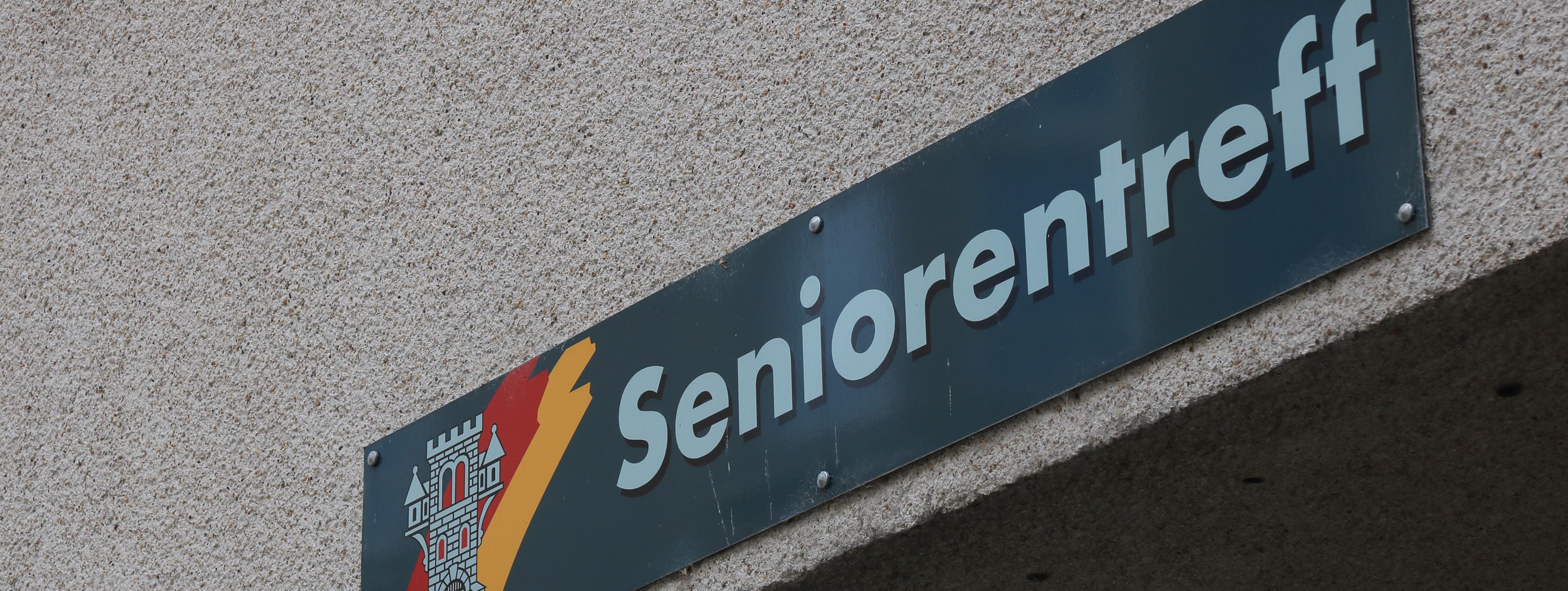 An der Hauswand des Treffs ist ein grünes Schild angebracht, auf dem in weißer Schrift "Seniorentreff" zu lesen ist. Auf dem Schild ist außerdem das Wappen der Stadt Menden zu sehen.