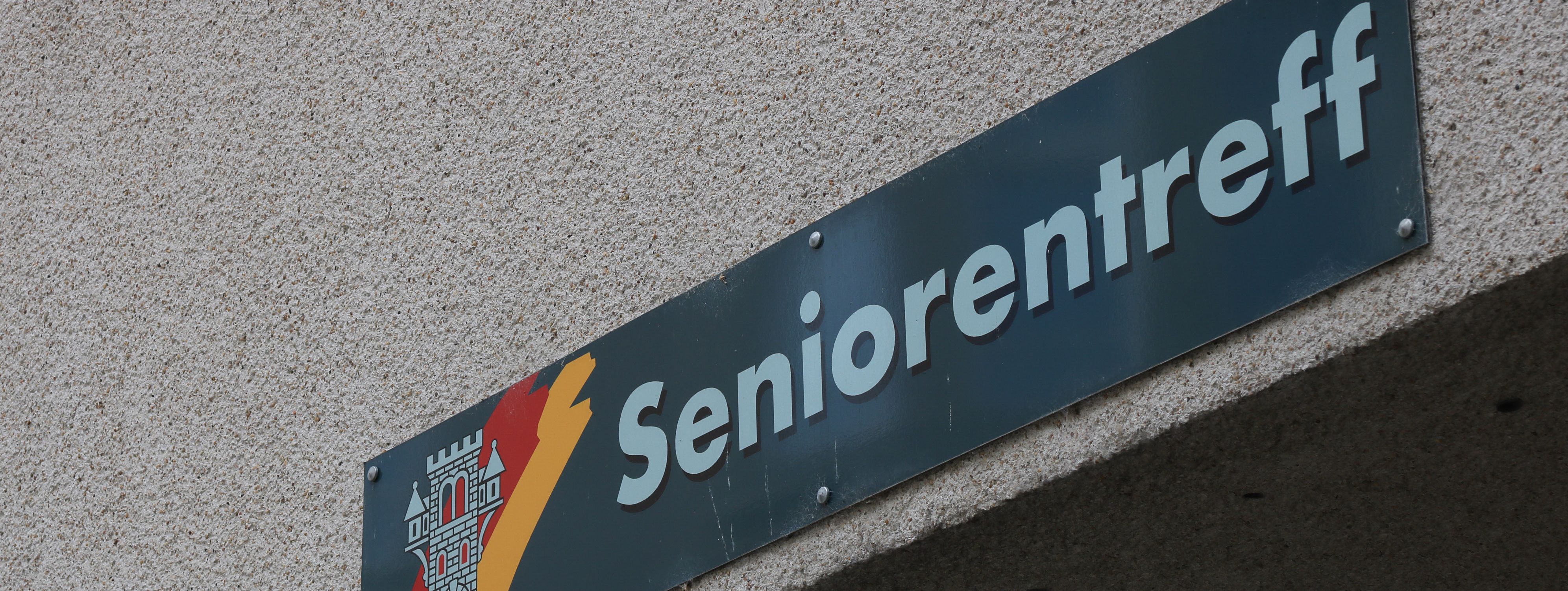 An der Hauswand des Treffs ist ein grünes Schild angebracht, auf dem in weißer Schrift "Seniorentreff" zu lesen ist. Auf dem Schild ist außerdem das Wappen der Stadt Menden zu sehen.