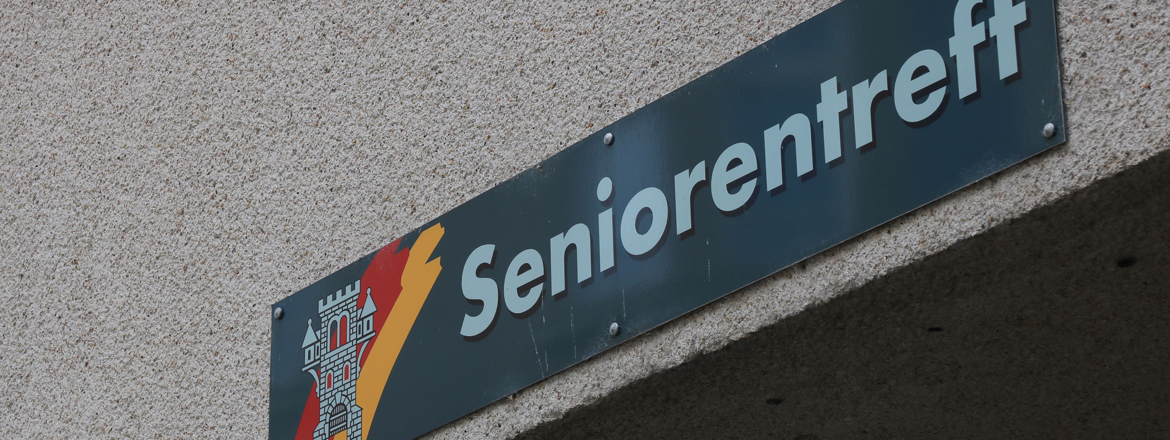 An der Hauswand des Treffs ist ein grünes Schild angebracht, auf dem in weißer Schrift "Seniorentreff" zu lesen ist. Auf dem Schild ist außerdem das Wappen der Stadt Menden zu sehen.