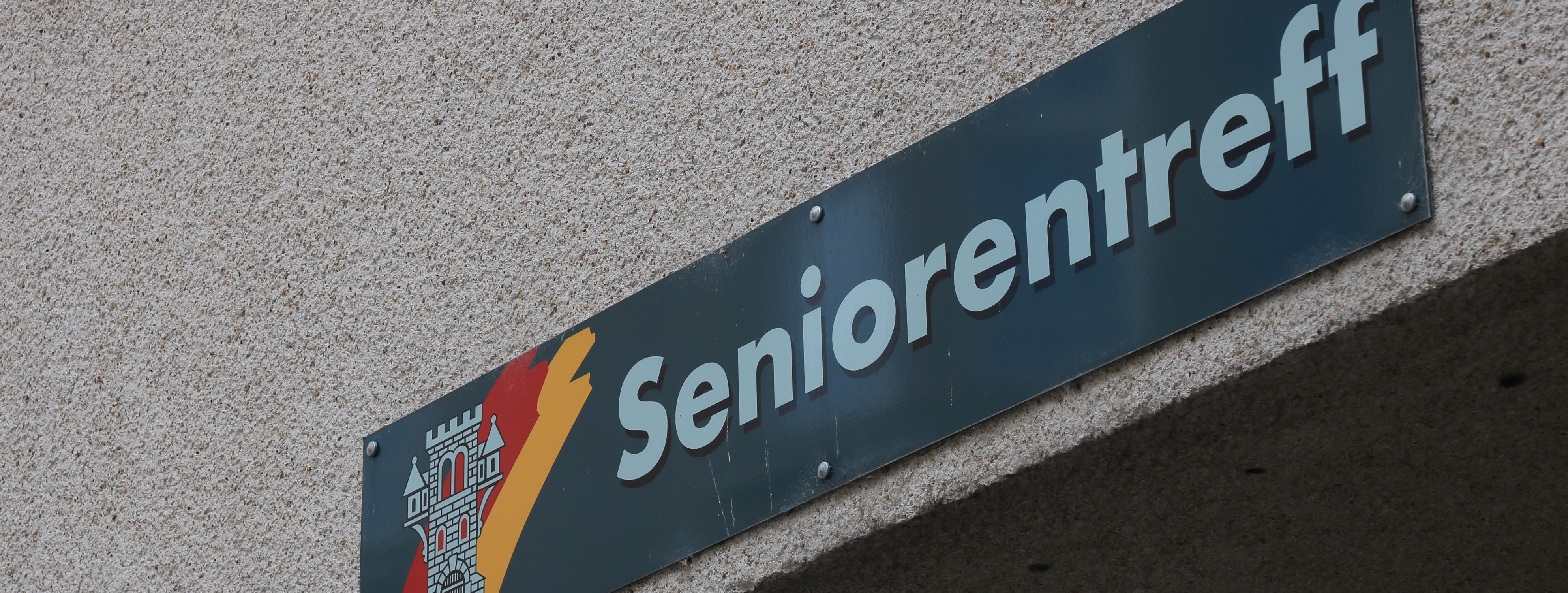 An der Hauswand des Treffs ist ein grünes Schild angebracht, auf dem in weißer Schrift "Seniorentreff" zu lesen ist. Auf dem Schild ist außerdem das Wappen der Stadt Menden zu sehen.