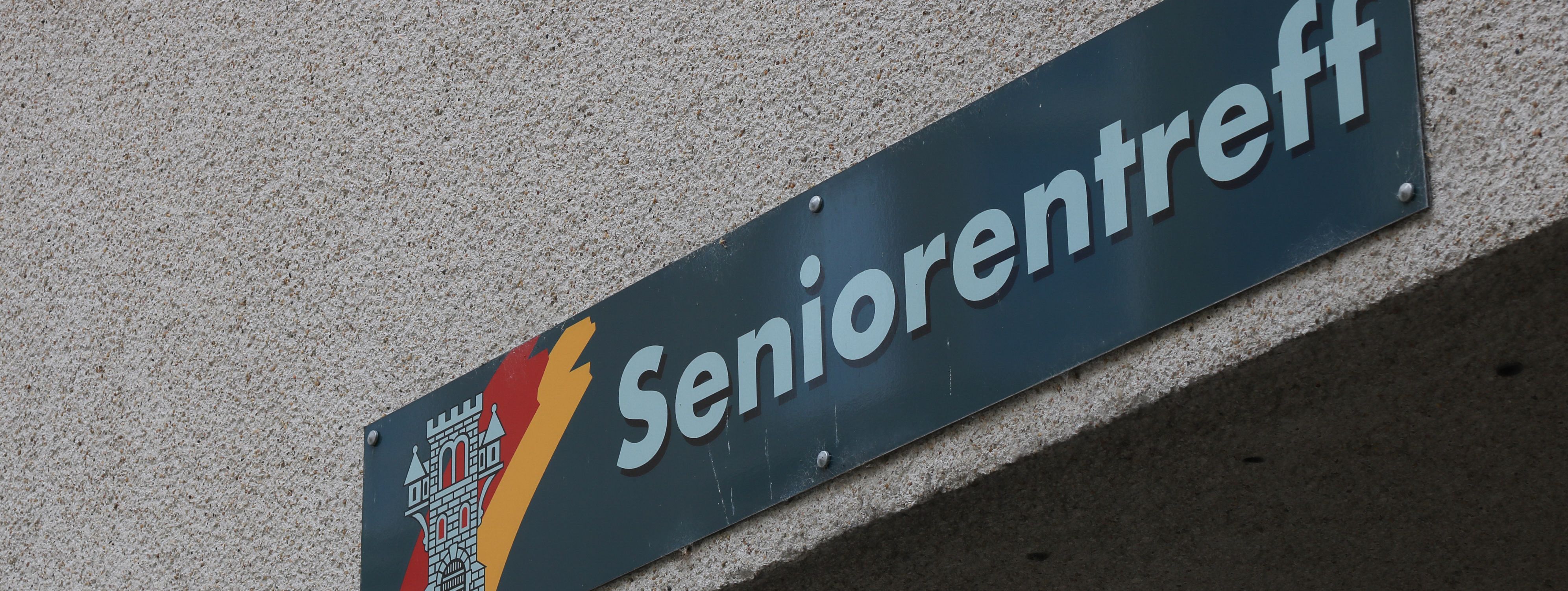 An der Hauswand des Treffs ist ein grünes Schild angebracht, auf dem in weißer Schrift "Seniorentreff" zu lesen ist. Auf dem Schild ist außerdem das Wappen der Stadt Menden zu sehen.