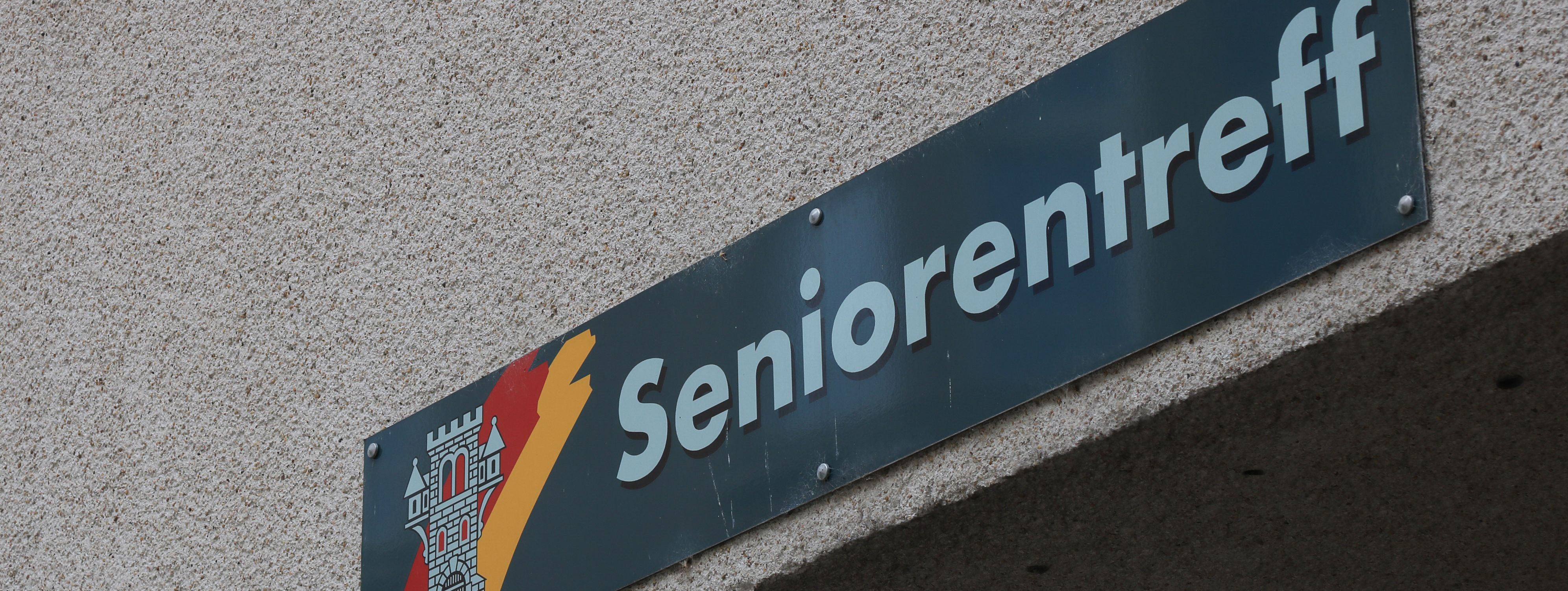 An der Hauswand des Treffs ist ein grünes Schild angebracht, auf dem in weißer Schrift "Seniorentreff" zu lesen ist. Auf dem Schild ist außerdem das Wappen der Stadt Menden zu sehen.