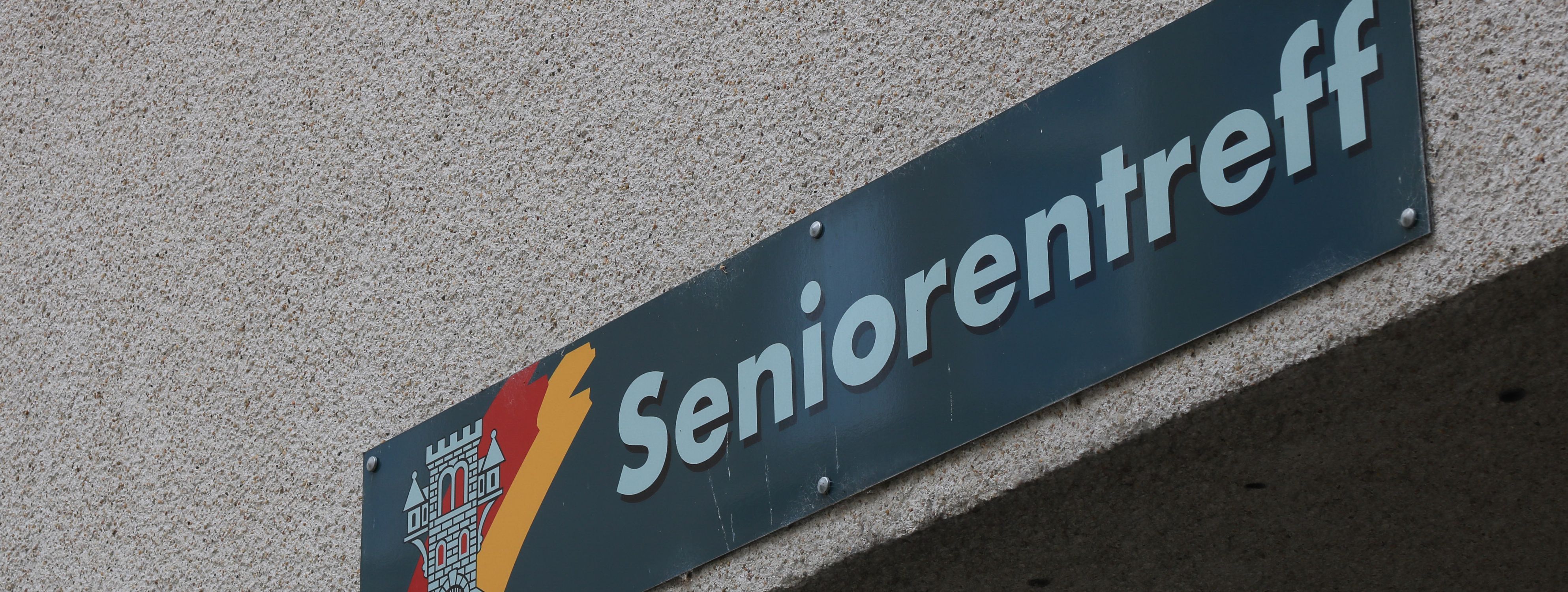 An der Hauswand des Treffs ist ein grünes Schild angebracht, auf dem in weißer Schrift "Seniorentreff" zu lesen ist. Auf dem Schild ist außerdem das Wappen der Stadt Menden zu sehen.