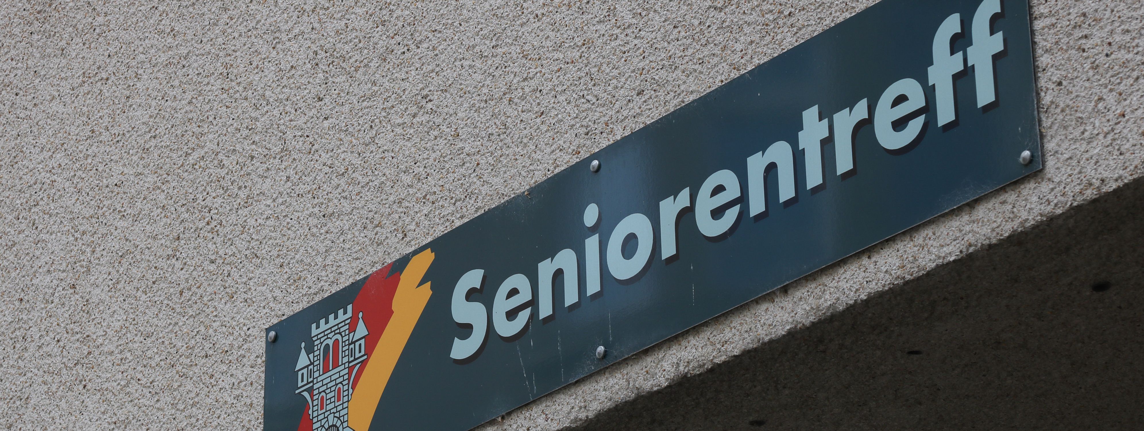 An der Hauswand des Treffs ist ein grünes Schild angebracht, auf dem in weißer Schrift "Seniorentreff" zu lesen ist. Auf dem Schild ist außerdem das Wappen der Stadt Menden zu sehen.