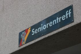 An der Hauswand des Treffs ist ein grünes Schild angebracht, auf dem in weißer Schrift "Seniorentreff" zu lesen ist. Auf dem Schild ist außerdem das Wappen der Stadt Menden zu sehen.