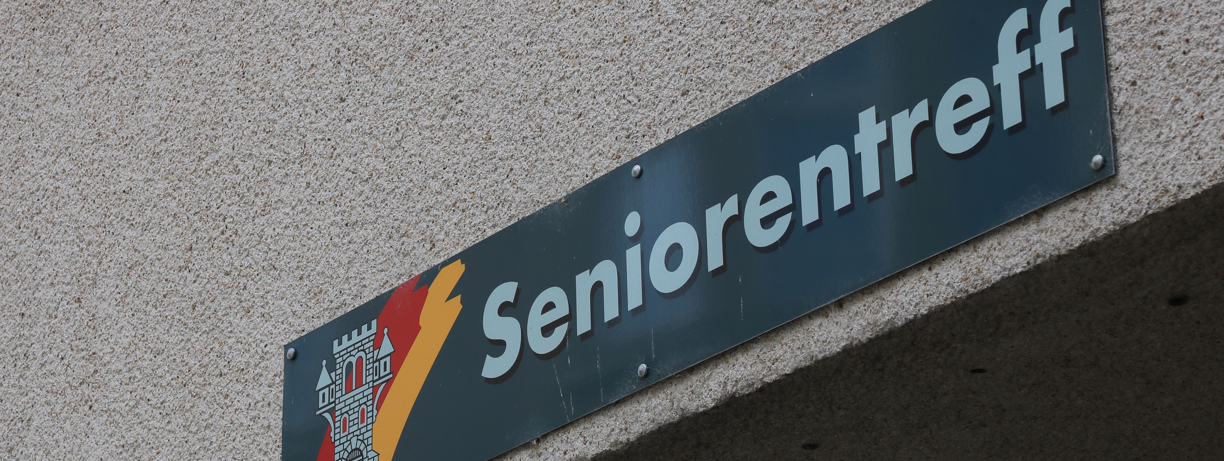 An der Hauswand des Treffs ist ein grünes Schild angebracht, auf dem in weißer Schrift "Seniorentreff" zu lesen ist. Auf dem Schild ist außerdem das Wappen der Stadt Menden zu sehen.