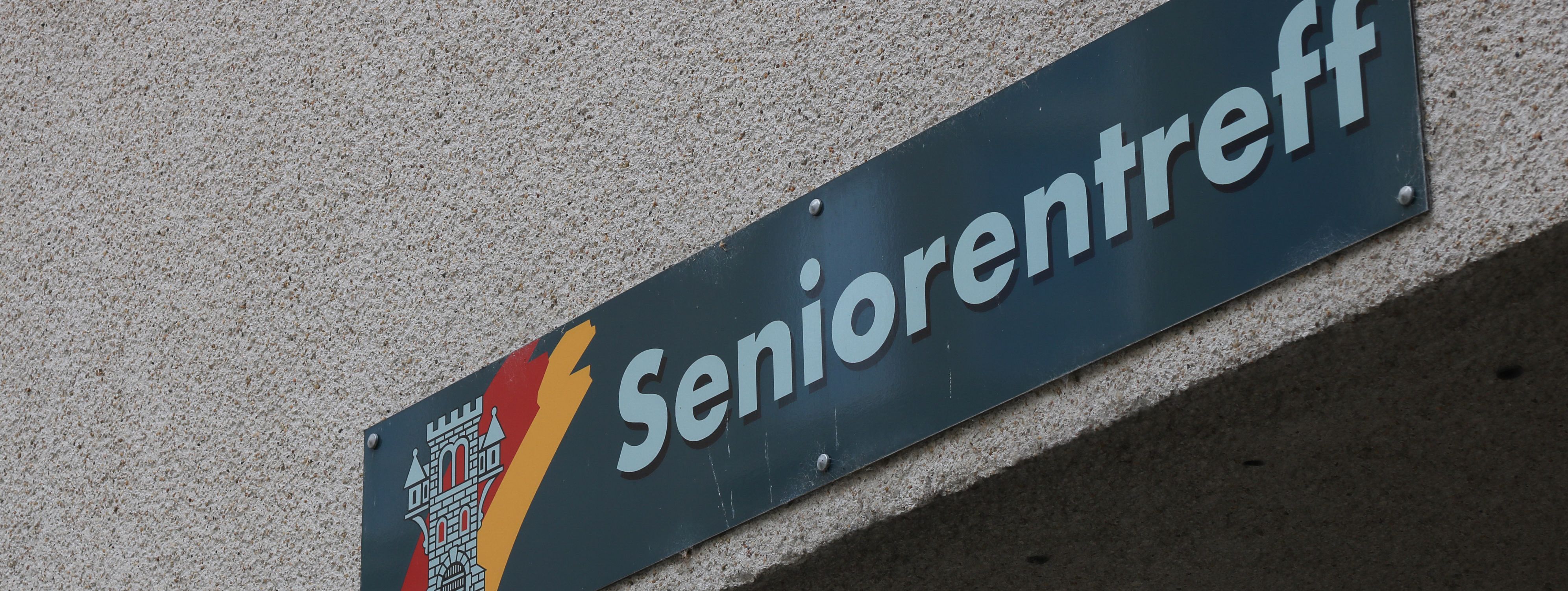 An der Hauswand des Treffs ist ein grünes Schild angebracht, auf dem in weißer Schrift "Seniorentreff" zu lesen ist. Auf dem Schild ist außerdem das Wappen der Stadt Menden zu sehen.