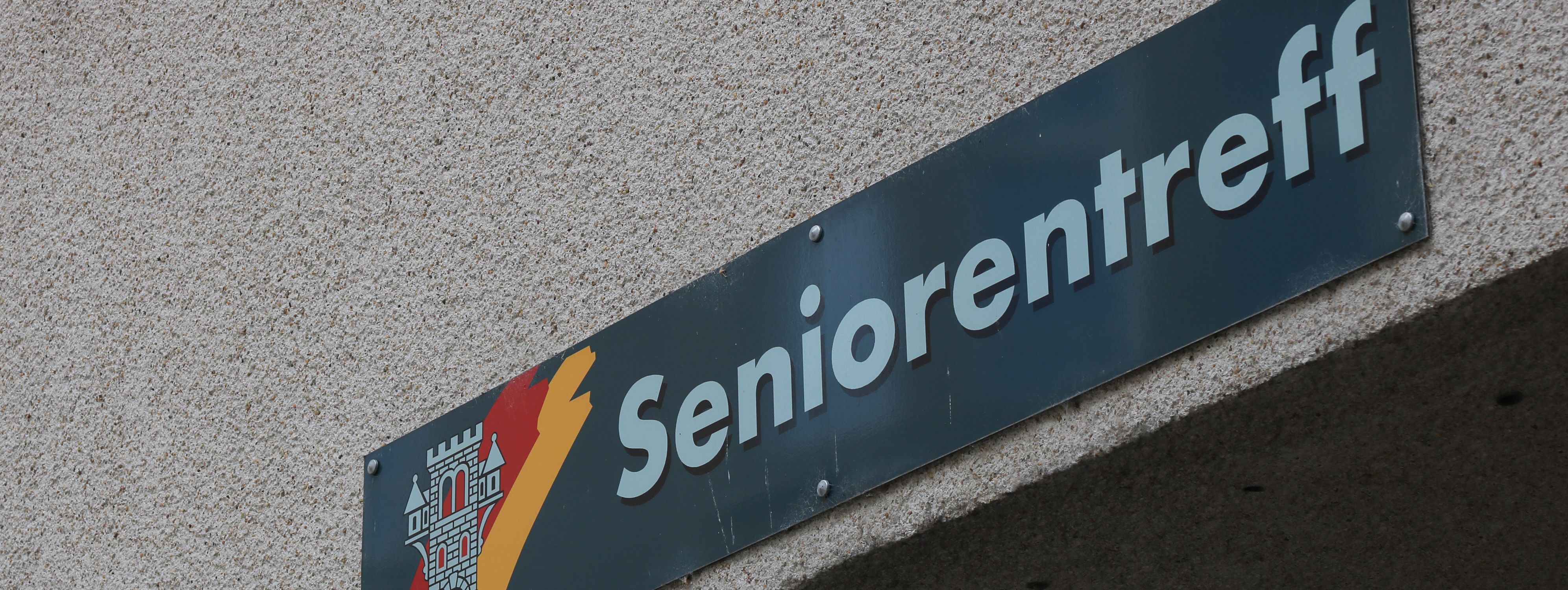 An der Hauswand des Treffs ist ein grünes Schild angebracht, auf dem in weißer Schrift "Seniorentreff" zu lesen ist. Auf dem Schild ist außerdem das Wappen der Stadt Menden zu sehen.