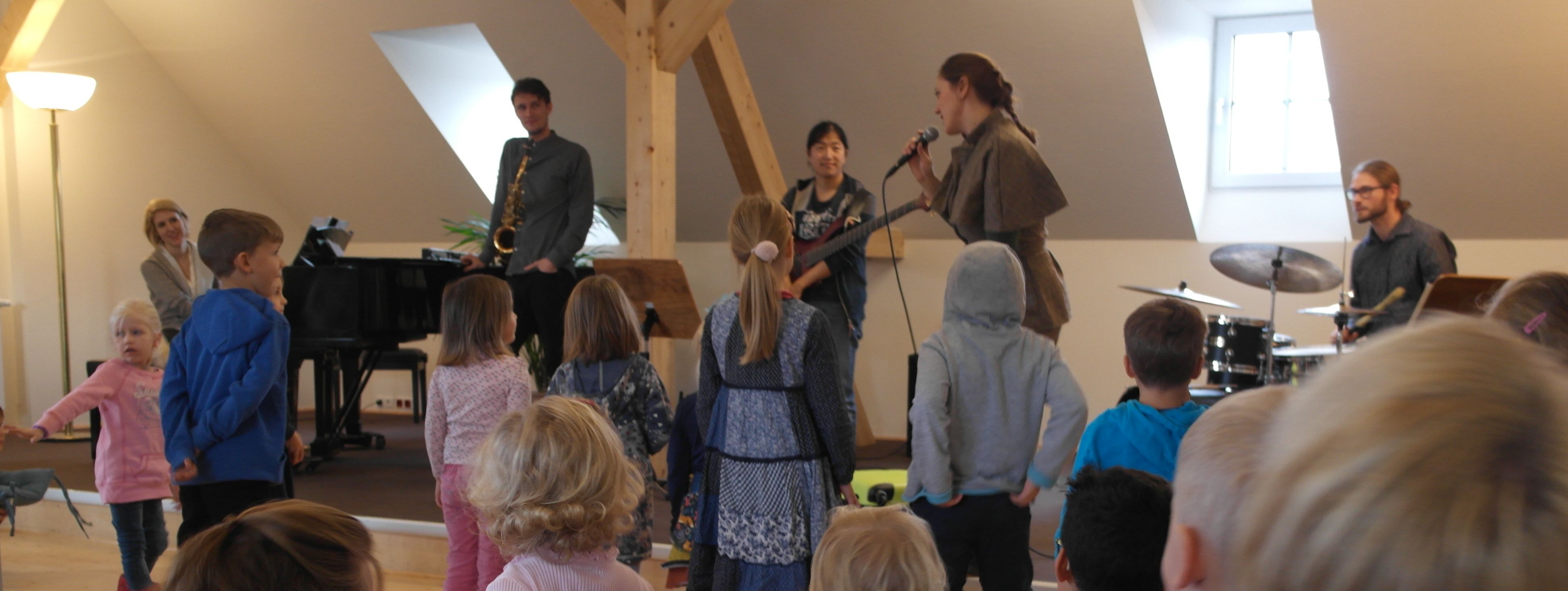 Vor der Bühne im Westflügel der Musikschule stehen Kinder und schauen, was die Musiker und "Hanna Fuchsfuß" auf der Bühne machen.