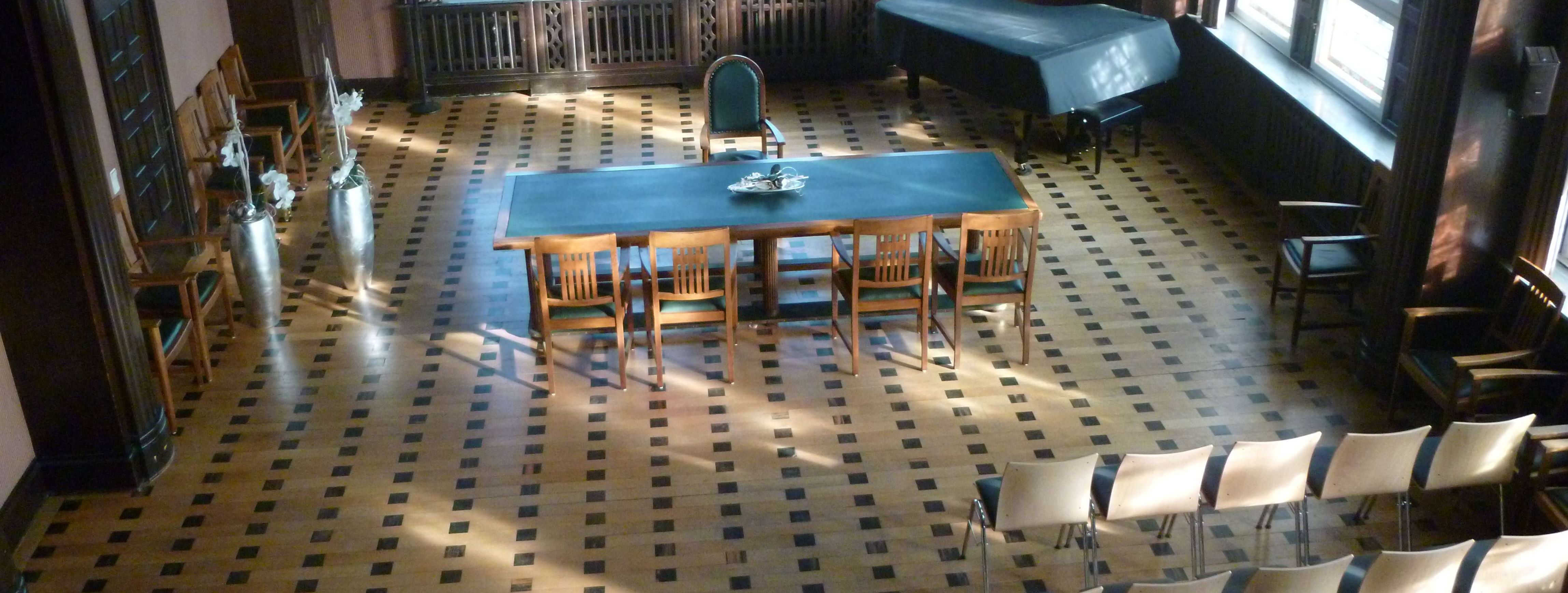 Historischer Ratssaal der Stadt Menden mit Bestuhlung, Blick von oben in den Saal, Buntglasfenster, Parkettboden, ein grüner Tisch steht quer, davor 4 Stühle. 