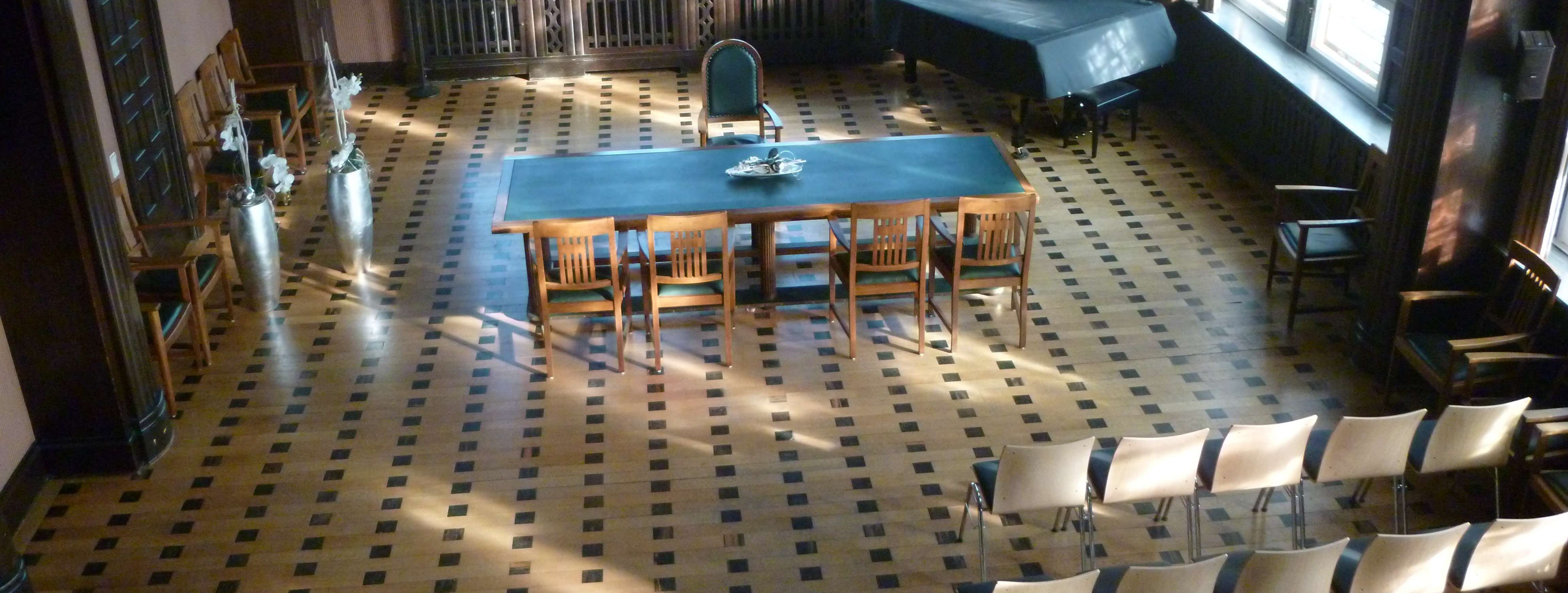 Historischer Ratssaal der Stadt Menden mit Bestuhlung, Blick von oben in den Saal, Buntglasfenster, Parkettboden, ein grüner Tisch steht quer, davor 4 Stühle. 