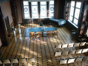 Historischer Ratssaal der Stadt Menden mit Bestuhlung, Blick von oben in den Saal, Buntglasfenster, Parkettboden, ein grüner Tisch steht quer, davor 4 Stühle. 