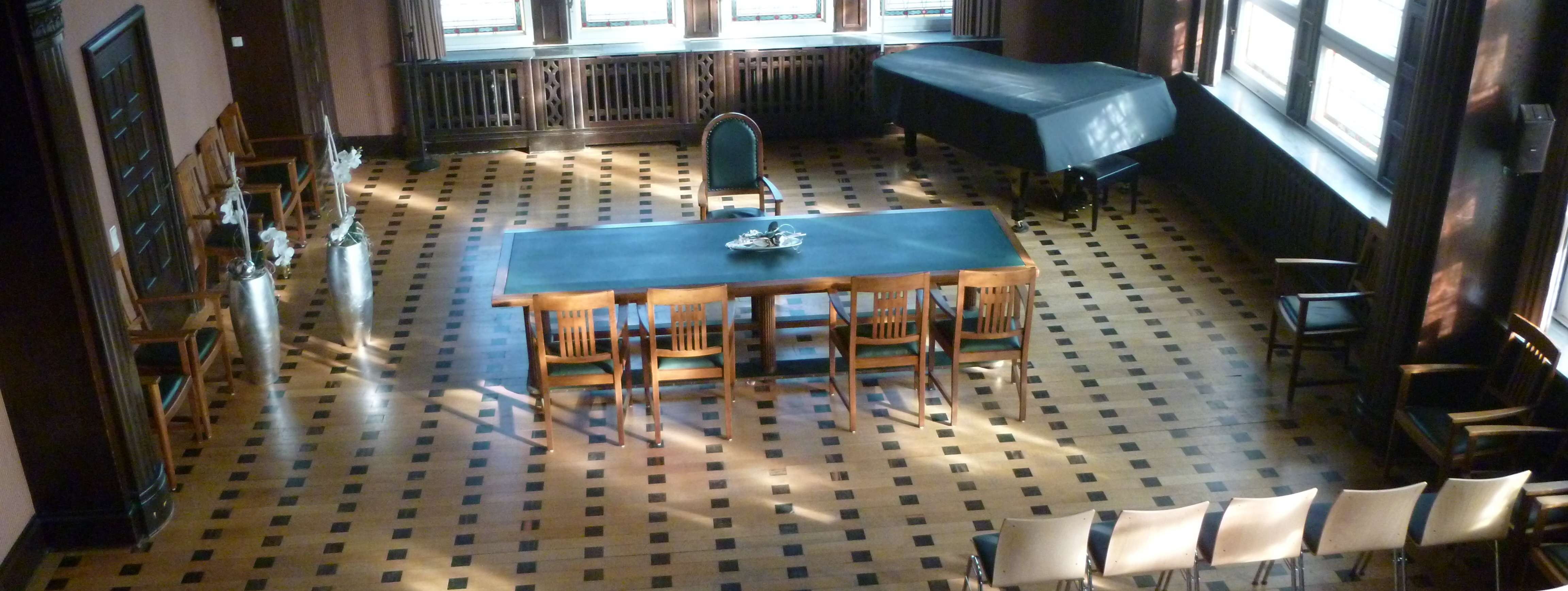 Historischer Ratssaal der Stadt Menden mit Bestuhlung, Blick von oben in den Saal, Buntglasfenster, Parkettboden, ein grüner Tisch steht quer, davor 4 Stühle. 