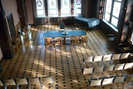 Historischer Ratssaal der Stadt Menden mit Bestuhlung, Blick von oben in den Saal, Buntglasfenster, Parkettboden, ein grüner Tisch steht quer, davor 4 Stühle. 