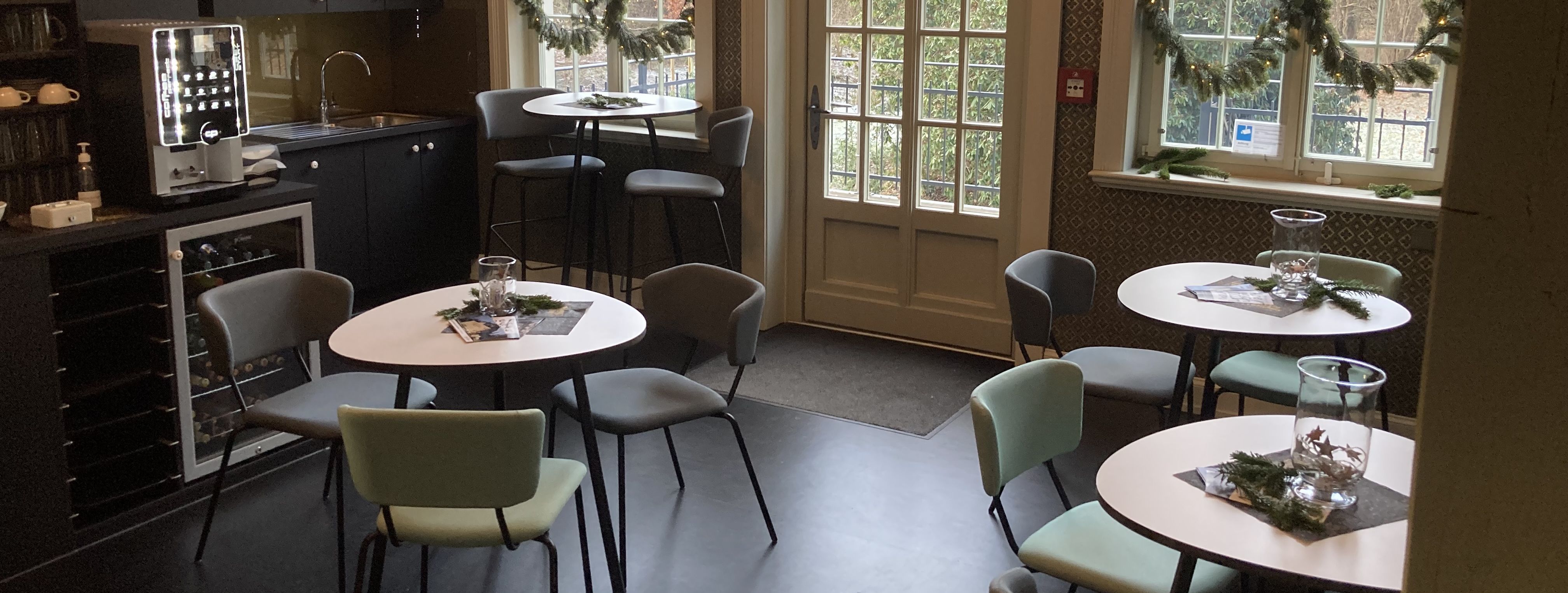 Blick in das winterlich geschmückte Museumscafé mit 3 kleinen Cafétischen, Kaffeebar und Parkzugang.. Der Raum hat gold-bläuliche Tapeten und einen schwarzen Linoleum-Boden. 