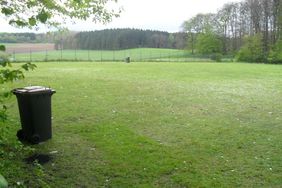 Auf dem grunen Rasen des Bolzplatzes sind noch deutlich zahllose kleine Plastik- und Papierfetzen zu sehen. Spuren der Feier von 1. Mai. Im Vordergrund steht eine Mülltonne.