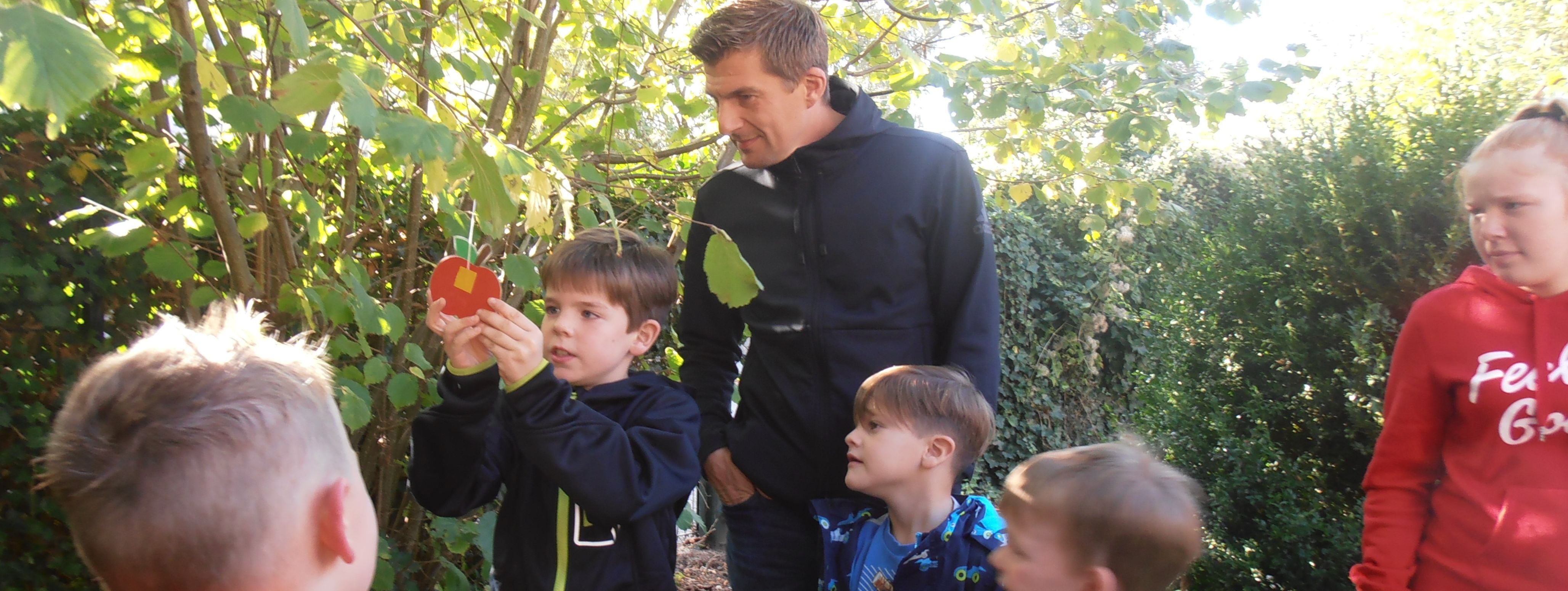 Vier Kinder und zwei Erwachsene stehen an einem Apfelbaum. Eines der Kinder hängt einen selbstgebastelten Apfel aus Pappe an einen Ast.