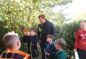 Vier Kinder und zwei Erwachsene stehen an einem Apfelbaum. Eines der Kinder hängt einen selbstgebastelten Apfel aus Pappe an einen Ast.