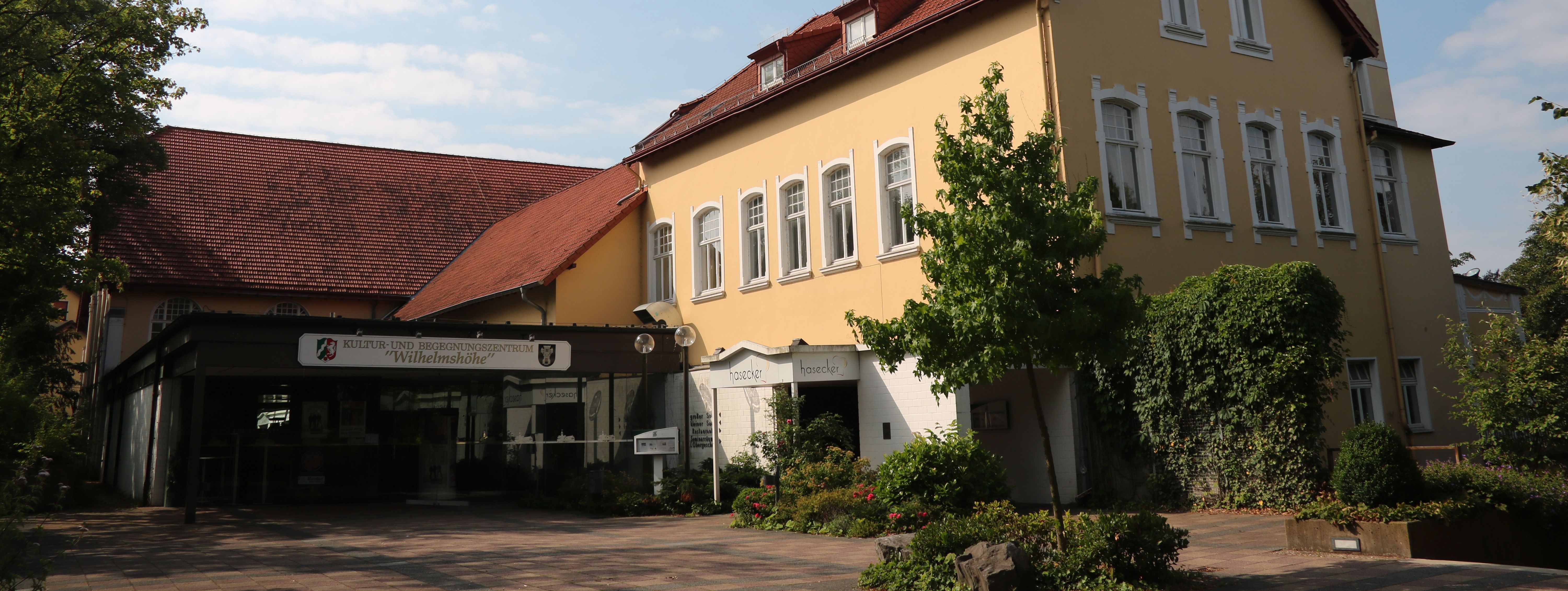 Der städtische Saalbetrieb: die Wilhelmshöhe Menden. Das Gebäude ist in hellem Gelb gestrichen, hat ein rotes Dach und Türmchen.