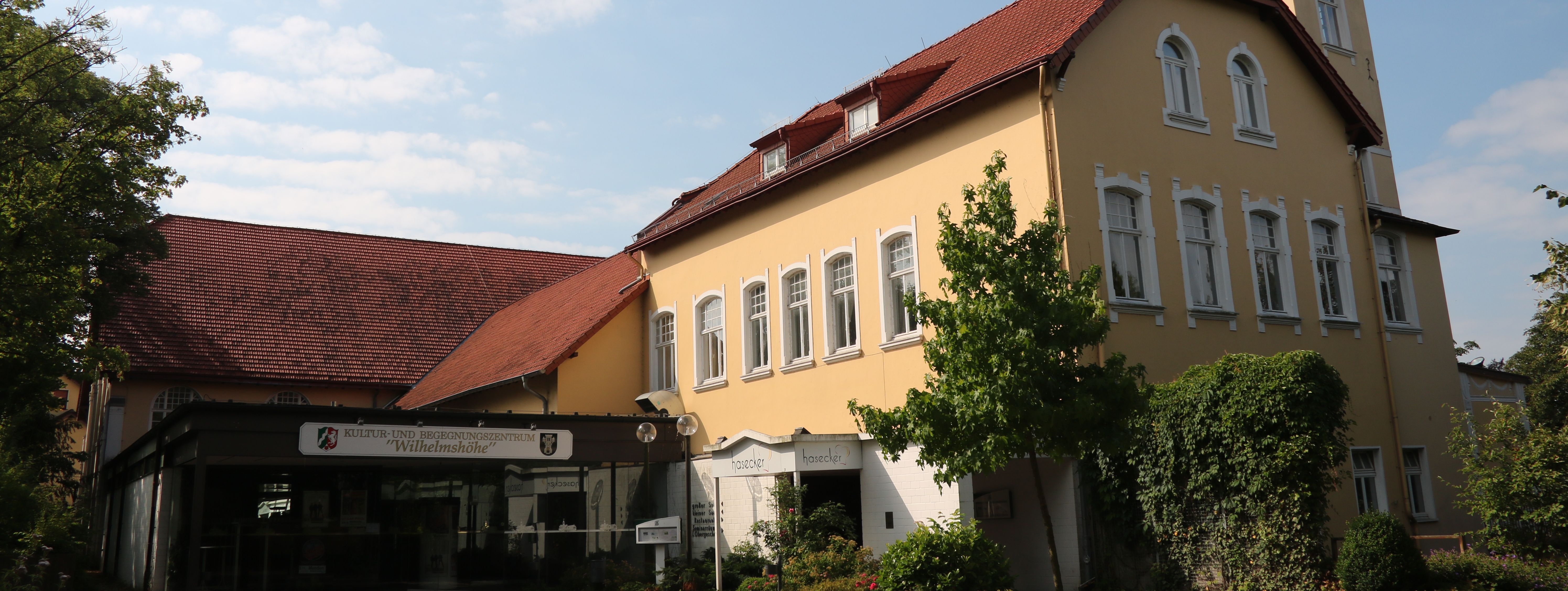 Der städtische Saalbetrieb: die Wilhelmshöhe Menden. Das Gebäude ist in hellem Gelb gestrichen, hat ein rotes Dach und Türmchen.