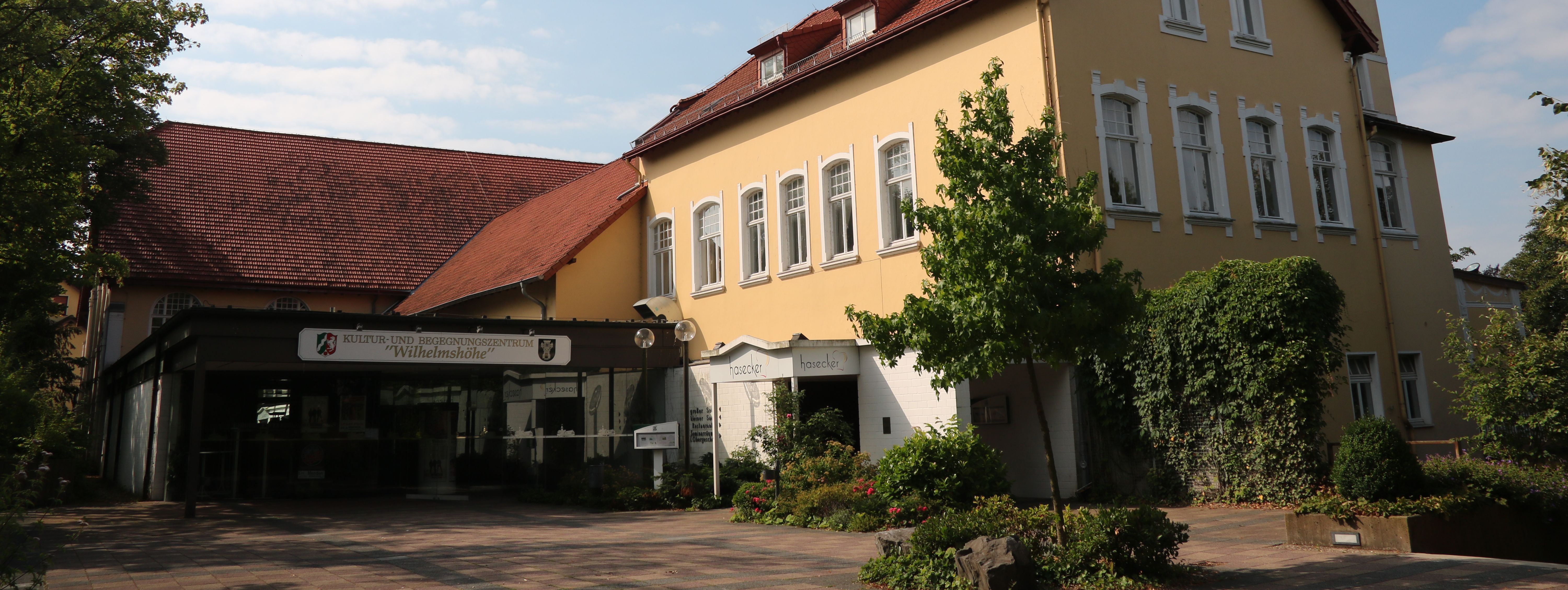 Der städtische Saalbetrieb: die Wilhelmshöhe Menden. Das Gebäude ist in hellem Gelb gestrichen, hat ein rotes Dach und Türmchen.