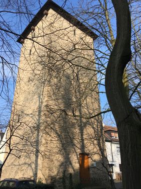 Ein Foto vom Poenigeturm. Linksseitig ist der Turm in Gänze mit Eingang. Rechtsseitig ist der Stamm eines Baumes. Der Turm ist von unten nach oben fotografiert vor blauem Himmel. 