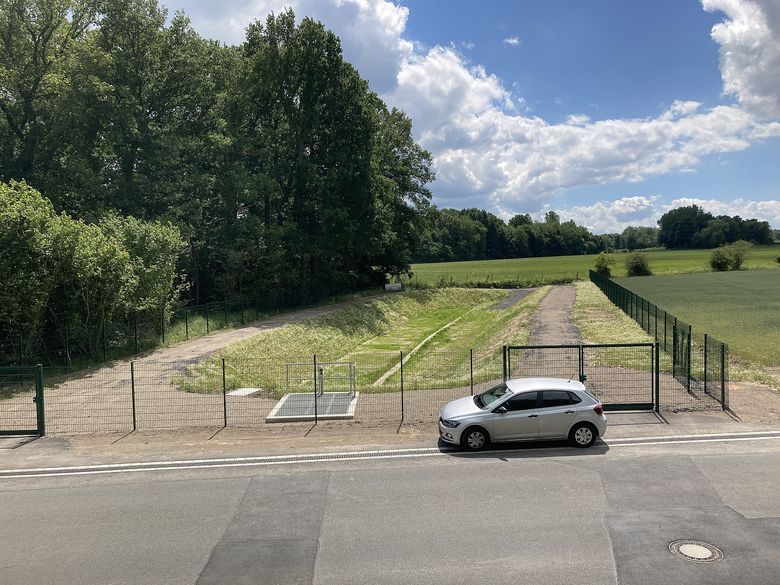 Das fertige Becken für den ersten Bauabschnitt Hämmer Süd mit einem Speichervolumen von rd. 470 cbm. Es ist rund 60 m lang, 10 - 15 m breit und maximal 1,20 m mit Wasser eingestaut. In der Mitte des Beckens wurde eine Sohle aus Betonhalbschalen angeordnet, um den Abfluss der Restwassermengen zu verbessern. Im Vordergrund ist eine größere Gitterrostabdeckung (ca. 3,70 x 2,70 m) angeordnet. Sie bedeckt den Drosselschacht, in dem der Abfluss aus dem Becken geregelt wird. Für den im Frühjahr 2021 anstehenden zweiten Bauabschnitt werden die dort benötigten Regenrückhaltebecken auf der Fläche im Hintergrund bis vor die Waldfläche in ca. 170 m Entfernung angeordnet.