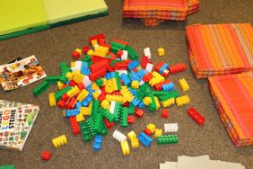 Auf dem braunen Teppich liegen die bunten Lego-Duplo-Steine, drumherum liegen Sitzkissen, auf denen die großen und kleinen Baumeister platznehmen können.