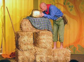 "Hans im Glück" steht gebäugt vor einem Stapel aus Strohballen und küsst ein großes, goldenes Ei, das ganz oben auf den Strohballen liegt.