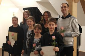 Die Musikschüler und ihre beiden Lehrer stehen vor einem aufgeklappten Flügel, einige halten ihre Urkunden und eine Rose in Händen.