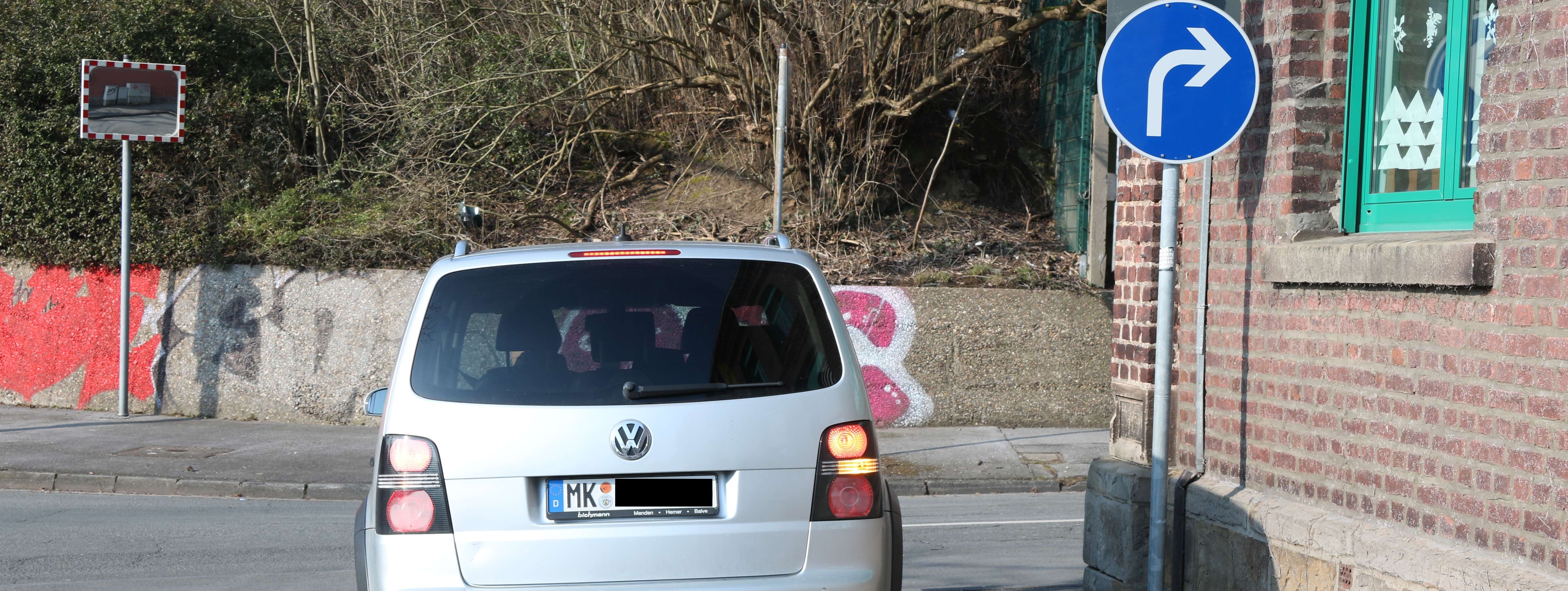 Ein silberner VW Touran fährt vom Parkplatz der Josefschule auf die Straße. Der Wagen hat den Blinker vorschriftsmäig nach rechts gesetzt. Neben dem Auto ist das neue Schild zu sehen. Es ist rund und zeigt einen weißen Pfeil auf blauem Grund. Es zeigt an, dass hier nur nach rechts abgebogen werden darf.