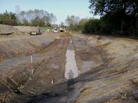 In einem ersten Bauabschnitt für das Gewerbegebiet Hämer Süd wurde u.a. ein Regenrückhaltebecken in Erdbauweise errichtet. Das Becken ist ca. 70 m lang, in der Sohle 8 - 9 m breit und im Mittel etwas über zwei Meter tief, das verfügbare Speichervolumen beträgt rund 475 Kubikmeter. In der Mitte des Beckens wird in Längsrichtung eine Betonrinne für die Entleerung des Beckens errichtet. Auf dem Bild ist das Kiesauflager für die Betonrinne gerade im Bau (Foto: Stadtentwässerung).