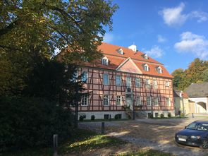 Das Foto zeigt die Frontansicht von Gut Rödinghausen. Ein imposantes Fachwerkhaus mit rosafarbenen Gefachen vor blauem Himmel. Ein herbstlicher Baum am linken Bildrand.