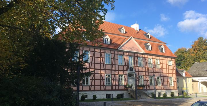 Das Foto zeigt die Frontansicht von Gut Rödinghausen. Ein imposantes Fachwerkhaus mit rosafarbenen Gefachen vor blauem Himmel. Ein herbstlicher Baum am linken Bildrand.