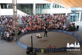 Unter dem Zeltdach vor dem neuen Rathaus ist eine Manege aufgebaut. Mitten in dieser Manege steht ein Zauberkünstler mit schwarzem Zylinderhut. Auf den Rängen sitzen zahlreiche Kinder mit ihren Eltern.