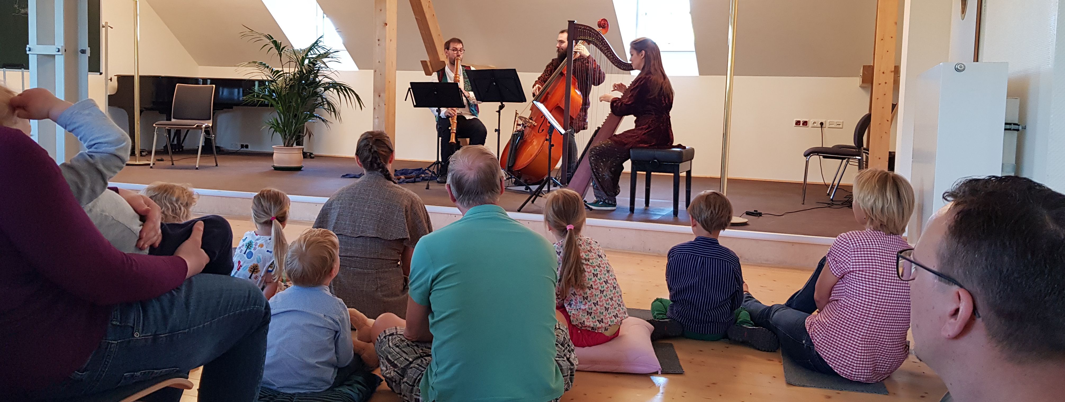 Kinder und Eltern sitzen auf dem Boden des Westflügels auf Kissen. Auf der Bühne sind drei Musiker zu sehen mit Harfe, Chello und Flöte.