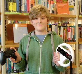 Der Schüler Jan Rüberg, Gaming-Experte der Stadtbücherei Menden, hält einen Controler in der einen und ein Lenkrad für ein Videospiel in der anderen Hand und lächelt verschmitzt in die Kamera