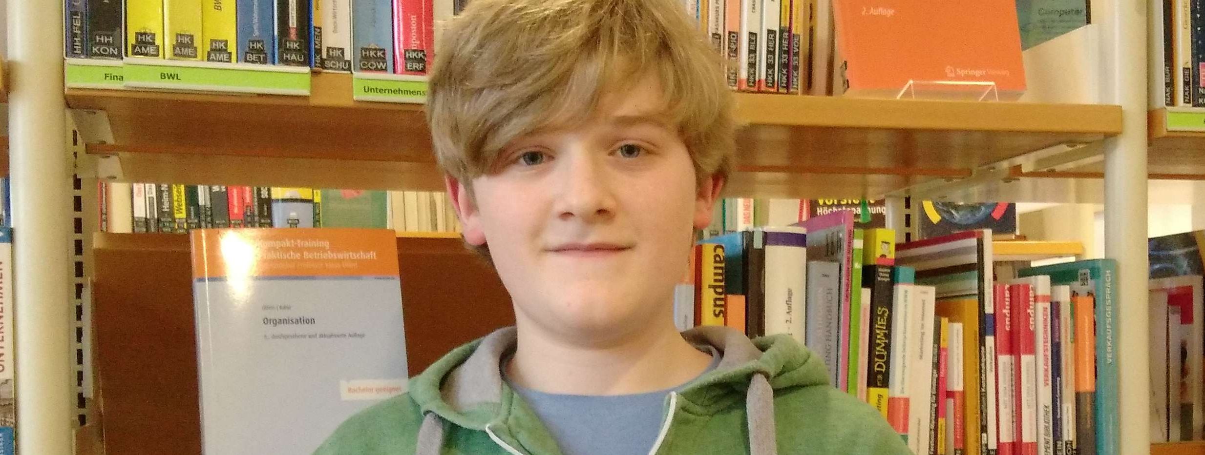 Der Schüler Jan Rüberg, Gaming-Experte der Stadtbücherei Menden, hält einen Controler in der einen und ein Lenkrad für ein Videospiel in der anderen Hand und lächelt verschmitzt in die Kamera