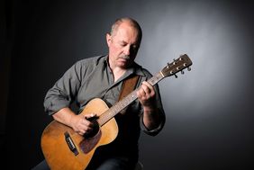 Sammy Vomacka steht vor einem grauen Hintergrund und hält seine Gitarre in Händen. Sein Blick ist nach Unten auf den Hals der Gitarre gerichtet.