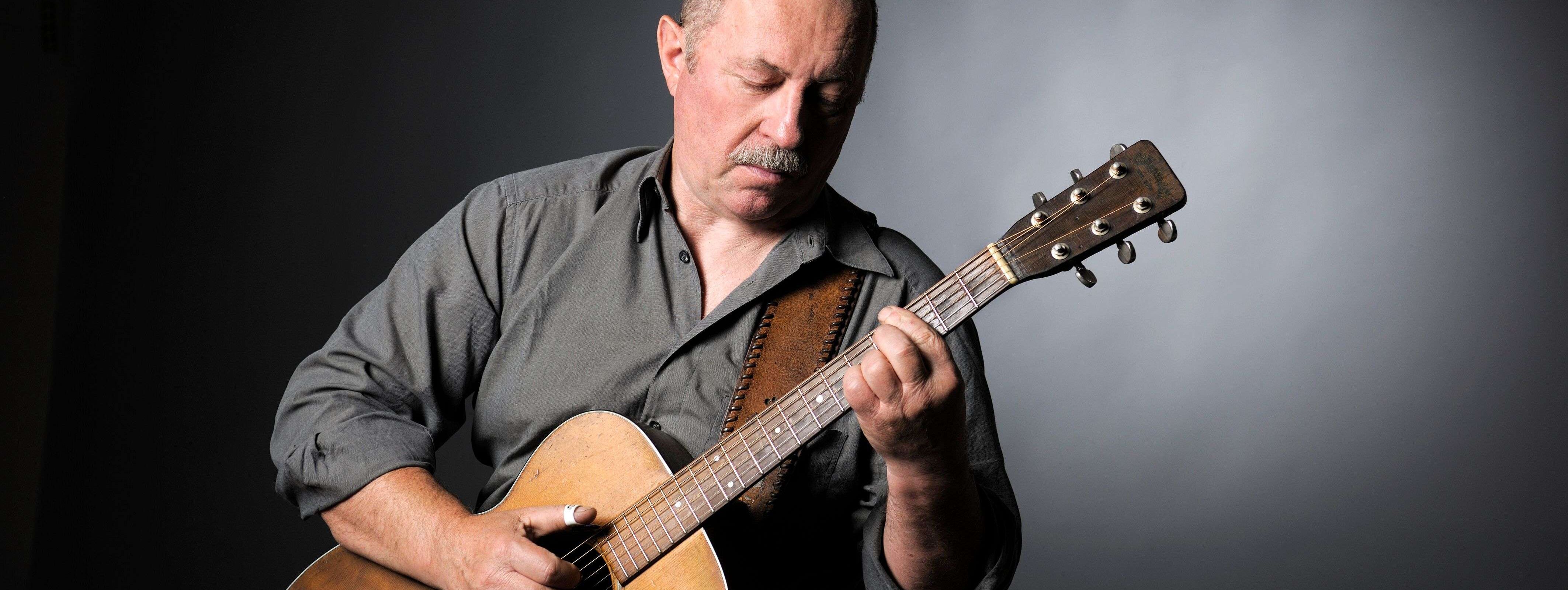 Sammy Vomacka steht vor einem grauen Hintergrund und hält seine Gitarre in Händen. Sein Blick ist nach Unten auf den Hals der Gitarre gerichtet.