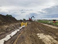 In der Planstraße E, östlich der Zufahrt zum Hof Riekenbrauck und parallel zur Hämmerstraße, werden Stahlbetonrohre mit 1,30 m Durchmesser verlegt. Zu sehen sind die fertig verlegten Rohre im Kanalgraben, der Rohrscheitel ist noch nicht abgedeckt. Die künftige Straßenfläche ist auf rund 12 m Breite freigelegt und der Oberboden abgeschoben. Im Hintergrund stehen zwei große Bagger, die mit der Verlegung des Regenwasserkanals beschäftigt sind.